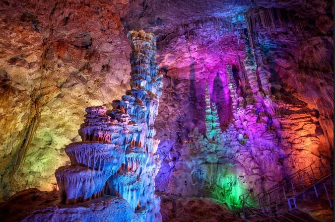 Beeindruckende Innenansicht der Canelobre-Höhlen bei Busot mit farbiger Beleuchtung, die Stalaktiten und Stalagmiten in leuchtenden Blau-, Lila- und Grüntönen hervorhebt.