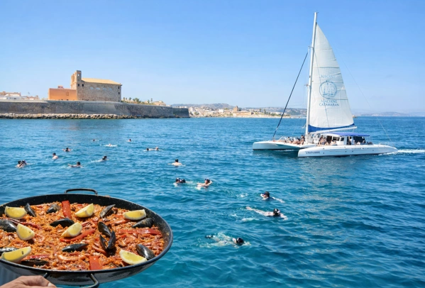 Paella an Bord eines Katamarans vor der Insel Tabarca, während Menschen im klaren Wasser schwimmen und ein Segelboot vor der Küste von Alicante fährt.