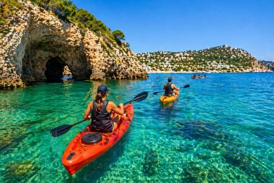 Personen paddeln mit Kajaks in kristallklarem Wasser vor einer Meeresgrotte an der Küste von Moraira (Alicante), umgeben von Felsenklippen und mediterraner Landschaft.