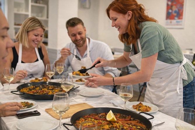 Gruppe von Teilnehmern genießt gemeinsam frisch zubereitete Paella während eines Kochkurses in Alicante, serviert von einer Person in Schürze in entspannter Atmosphäre.