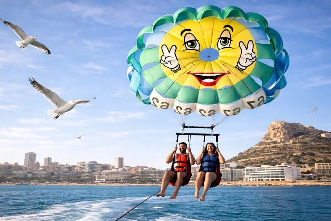 Zwei Touristen beim Parasailing in Alicante über dem Meer mit einem bunten Fallschirm, während Möwen um sie herum fliegen und die Burg Santa Bárbara im Hintergrund an der Küste zu sehen ist.