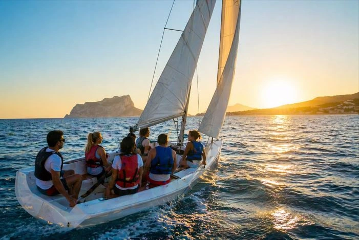 Gruppe von Personen segelt bei Sonnenuntergang auf einem kleinen Segelboot vor der Küste von Moraira (Alicante) und genießt das Meer sowie die Ausblicke auf die Costa Blanca.
