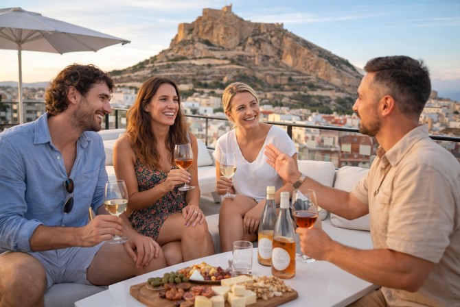 Kleine Gruppe von Touristen genießt eine Weinprobe auf einer Dachterrasse in Alicante mit Blick auf die Burg Santa Bárbara bei Sonnenuntergang.