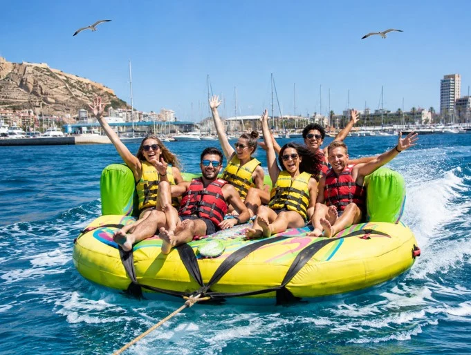 Gruppe von Touristen auf einem aufblasbaren Crazy Sofa, das über das Meer vor Alicante gezogen wird, mit Schwimmwesten, fliegenden Möwen und dem Hafen im Hintergrund.