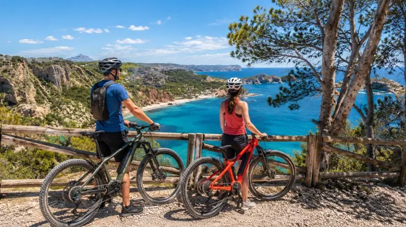 Zwei Radfahrer mit E-Bikes an einem Aussichtspunkt in Jávea mit Blick auf die Küste, türkisblaues Meer und mediterrane Landschaft.