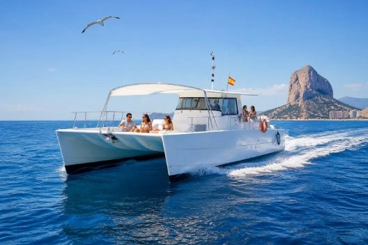 Katamaran mit Touristen vor der Küste von Calpe, mit blauem Mittelmeer, Möwen am Himmel und dem Peñón de Ifach im Hintergrund.