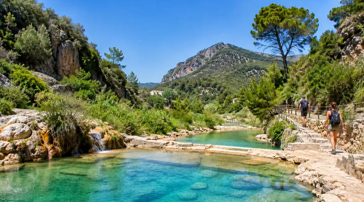Wanderer auf einem Weg neben den natürlichen Badebecken der Fuente de los Baños in Montanejos mit türkisfarbenem Wasser, Bergen und mediterraner Natur.