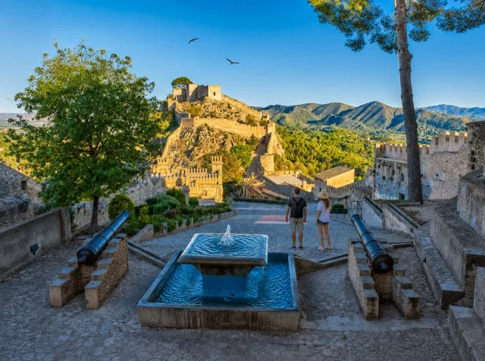 Blick auf die Burg von Xàtiva mit Brunnen, historischen Mauern, Bergen und zwei Touristen an einem Aussichtspunkt.