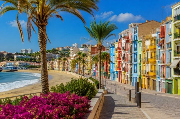Bunte Häuser direkt am Meer in Villajoyosa mit Strandpromenade, Palmen und Strand an einem sonnigen Tag.
