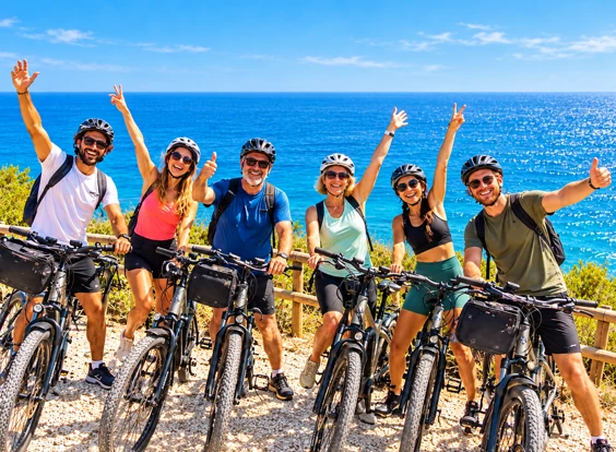Gruppe von Touristen mit E-Bikes, die an einem Aussichtspunkt am Meer jubeln, mit blauem Himmel und offenem Mittelmeer im Hintergrund.