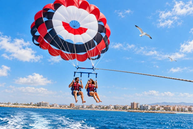 Zwei Touristen beim Parasailing über dem Mittelmeer in Torrevieja, mit der Küste im Hintergrund, blauem Himmel, Wolken und Möwen.
